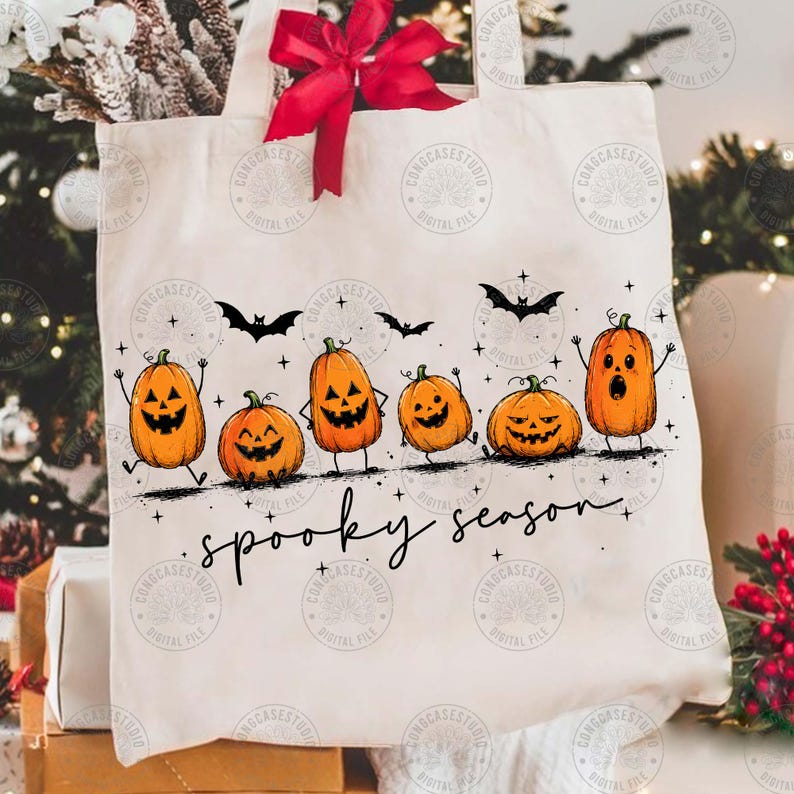 May include: A cream-colored canvas tote bag featuring a red ribbon tied at the top handle. The front displays a graphic of six orange jack-o'-lanterns with various facial expressions, black bats, and the handwritten black text spooky season.