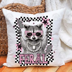 May include: White square pillow with a raccoon graphic wearing pink sunglasses and a pearl necklace. The raccoon is framed by a black and white checkered border with pink stars and the word "FERAL" in pink block letters.