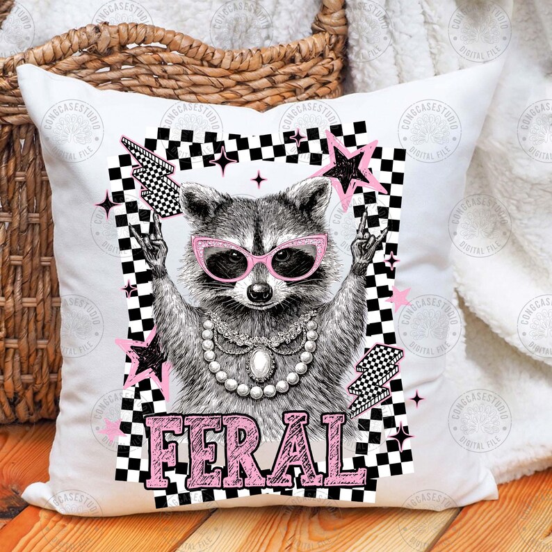 May include: White square pillow with a raccoon graphic wearing pink sunglasses and a pearl necklace. The raccoon is framed by a black and white checkered border with pink stars and the word "FERAL" in pink block letters.