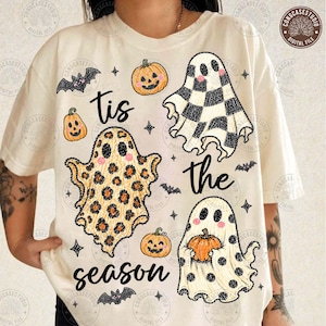 May include: A cream-coloured t-shirt featuring a graphic print with the text 'tis the season'. The design includes three ghosts with different patterns: checkered, leopard print, and polka dot, alongside orange pumpkins and small black bats.