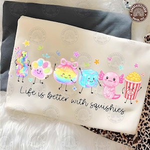 Puede incluir: Una camiseta color crema con un gráfico colorido de seis personajes de dibujos animados que incluyen una barra de mantequilla, una pata, una bolsa, un cubo de hielo, un ajolote y un cubo de palomitas, con el texto Life is better with squishies.