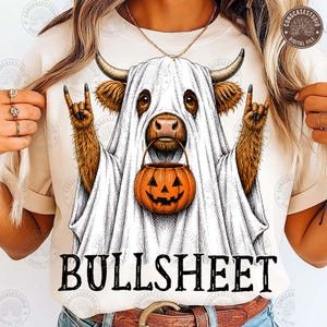 Può includere: Una t-shirt color crema con la grafica di una mucca Highland marrone che indossa un costume da fantasma bianco. La mucca fa gesti rock-and-roll con le mani e tiene in mano un secchiello a forma di zucca arancione. Il testo BULLSHEET è stampato sotto.