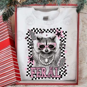 May include: White sweatshirt featuring a raccoon wearing pink sunglasses and a pearl necklace, with the word "FERAL" in pink. The design includes a black and white checkered background, pink stars, and lightning bolts.