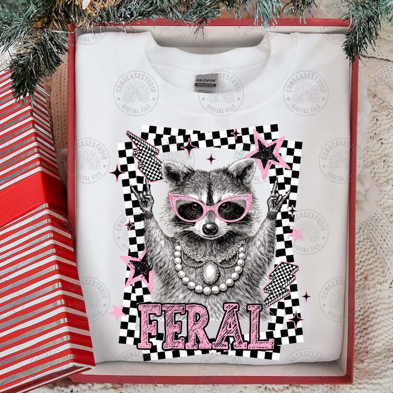 May include: White sweatshirt featuring a raccoon wearing pink sunglasses and a pearl necklace, with the word "FERAL" in pink. The design includes a black and white checkered background, pink stars, and lightning bolts.