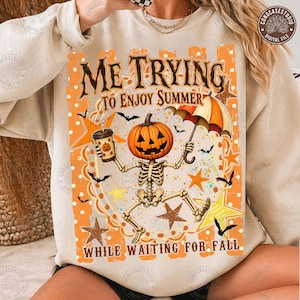 Pode incluir: Uma camisola bege com um gráfico de um esqueleto com cabeça de abóbora segurando uma chávena de café e um guarda-chuva. O texto diz ME TRYING TO ENJOY SUMMER WHILE WAITING FOR FALL. O design inclui estrelas cor de laranja, morcegos e pontos.