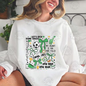 Puede incluir: Sudadera blanca con un dise&ntilde;o tem&aacute;tico de f&uacute;tbol. El dise&ntilde;o incluye un bal&oacute;n de f&uacute;tbol, lazos verdes, una bota de f&uacute;tbol y las frases "Let's Kick Some Grass", "Game Day" y "One Shot One Goal."