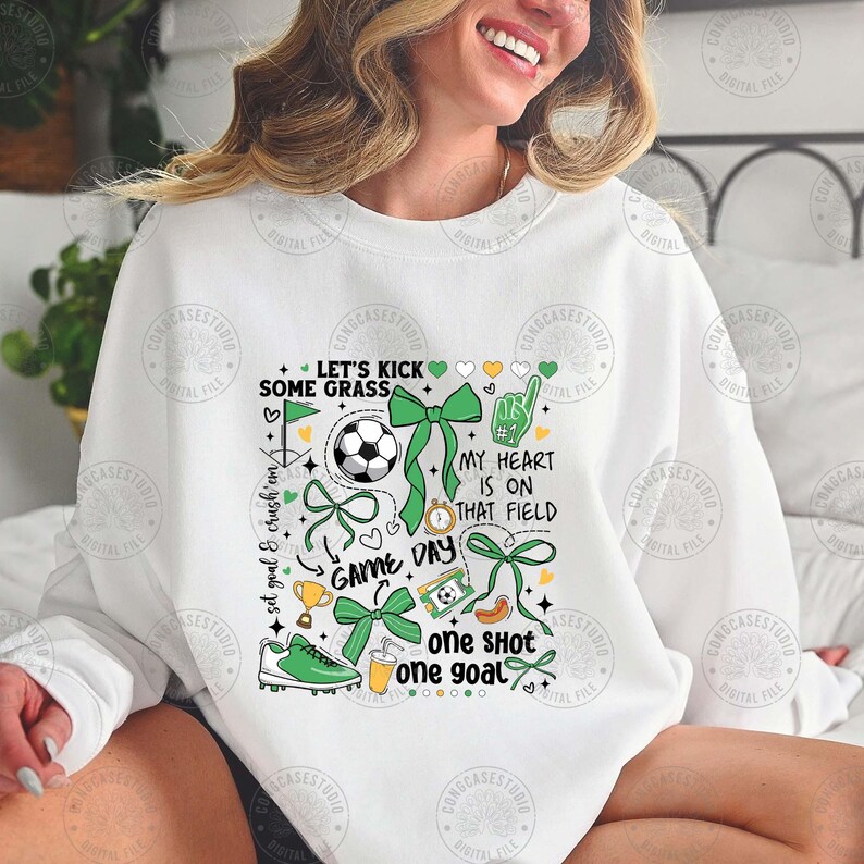 Puede incluir: Sudadera blanca con un dise&ntilde;o tem&aacute;tico de f&uacute;tbol. El dise&ntilde;o incluye un bal&oacute;n de f&uacute;tbol, lazos verdes, una bota de f&uacute;tbol y las frases "Let's Kick Some Grass", "Game Day" y "One Shot One Goal."