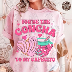 May include: Pink t-shirt with the text "YOU'RE THE CONCHA TO MY CAFECITO" in pink and white. The graphic includes a pink concha pastry and a pink mug with a drink and a straw. Hearts and stars are also included in the design.