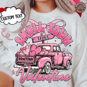 May include: A white sweatshirt with a pink vintage truck graphic, filled with heart-shaped balloons. The design includes the words "Small Town Valentine" and "Hometown". A cloud-shaped text box reads "CUSTOM TEXT".