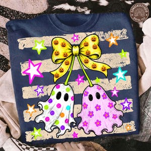 May include: A navy blue t-shirt featuring a graphic of two ghosts hanging from a large yellow bow patterned with orange pumpkins. One ghost is white with colorful polka dots, and the other is light purple with pink flowers, surrounded by stars.