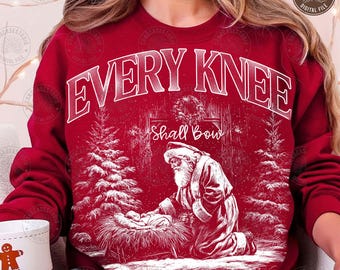 May include: A red sweatshirt with a white graphic featuring the text "EVERY KNEE SHALL BOW" above an illustration of Santa Claus kneeling before baby Jesus. The design includes Christmas trees and the word "Philippiar".