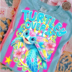 May include: A light blue t-shirt with the phrase "TURTLES OVER IT" in a playful font. The graphic features a colorful cartoon turtle with pink and blue accents, surrounded by flowers and stars. The shirt is made of a soft material.