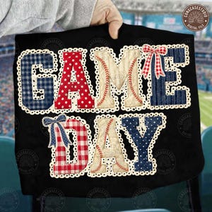 May include: Black t-shirt with the words "GAME DAY" in a patchwork design. The letters are in red polka dots, blue gingham, and baseball-themed beige with red stitching. White lace trim surrounds each letter, with bows on the letters E and D.