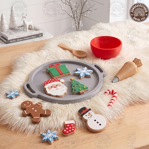 May include: A wooden toy set featuring a gray circular tray with handles, holding five Christmas-themed cookies shaped like a gift, snowflake, Santa Claus, Christmas tree, and snowman. Includes a red bowl, wooden spoon, piping bag, and candy cane.