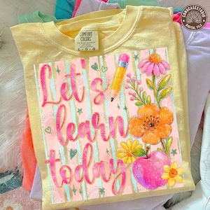 May include: A pale yellow t-shirt featuring a graphic print with the text "Let's learn today" in pink cursive script. The design includes a yellow pencil, a pink apple, and various orange, pink, and yellow flowers on a striped background.