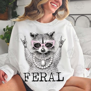 May include: White sweatshirt featuring a raccoon wearing pink sunglasses and a pearl necklace, with the word "FERAL" in bold black letters. The raccoon is making a rock and roll hand gesture.