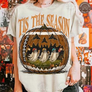 May include: A cream-colored t-shirt featuring a large graphic of a carved pumpkin shaped like a football. Inside the pumpkin, three ghosts wearing football helmets play on a field. The text 'TIS THE SEASON' is printed in orange above the pumpkin.