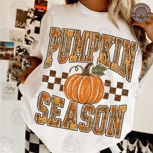 May include: A white t-shirt featuring the text PUMPKIN SEASON in large block letters with a camouflage and autumn leaf pattern. A central orange pumpkin illustration sits between the words, accompanied by a brown and white checkered pattern.