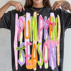 May include: A charcoal gray t-shirt featuring the text PUMPKIN SEASON in large, colorful, brushstroke-style letters. The design includes illustrations of pumpkins in shades of purple, pink, and orange, accented with bright green and pink bows.