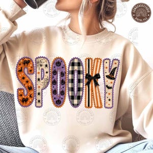 May include: A beige long-sleeve sweatshirt featuring the word SPOOKY in large, patterned letters. Each letter has a unique design including orange cats, spiderwebs, purple pumpkins, black and white plaid, stripes with a black bow, and a witch hat.
