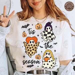 Puede incluir: Una camiseta blanca con un estampado gráfico que incluye el texto 'tis the season' rodeado de tres fantasmas con diferentes patrones, incluyendo estampado de leopardo, cuadros blancos y negros, y lunares, junto a pequeñas calabazas naranjas y murciélagos.