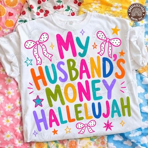 K&ouml;nnte beinhalten: Ein wei&szlig;es T-Shirt mit dem bunten Schriftzug My Husband's Money Hallelujah in Pink, T&uuml;rkis, Orange, Gr&uuml;n und Lila. Das Design ist mit wei&szlig;-schwarzen gepunkteten Schleifen und verschiedenen kleinen gelben, orangen und t&uuml;rkisen Sternen verziert.