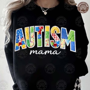 以下が含まれることがあります： カラフルなパッチワーク文字で「AUTISM」と白い筆記体で「mama」と書かれた黒いスウェットシャツ。文字は、水玉模様、花柄、パズルピースなど、さまざまなパターンで作られています。