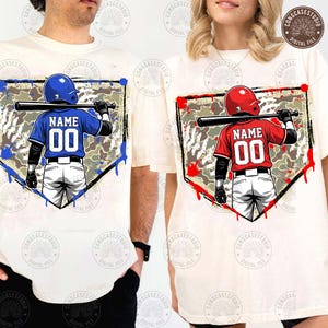 May include: Two cream-coloured t-shirts with baseball-themed graphics. One shirt features a blue baseball player graphic, the other a red one. Each graphic includes the word "NAME" and the number "00". The graphics are set within a home plate shape.