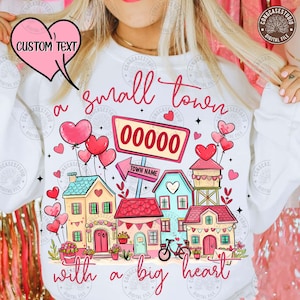 May include: White sweatshirt with a colorful graphic of a small town, featuring houses, hearts, and a bicycle. The text reads "a small town with a big heart" and includes space for custom text and a zip code.