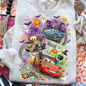 May include: A light beige sweatshirt featuring a colorful graphic print of three cartoon cars decorated for Halloween. The design includes a red race car, a brown tow truck, and a blue car wearing a witch hat, surrounded by ghosts, pumpkins, bats, and candy.