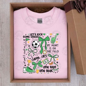 Puede incluir: Sudadera rosa claro con un dise&ntilde;o tem&aacute;tico de f&uacute;tbol. El dise&ntilde;o incluye un bal&oacute;n de f&uacute;tbol, lazos verdes y el texto "Let's kick some grass", "Game Day" y "My heart is on that field". Presentada en una caja de regalo.