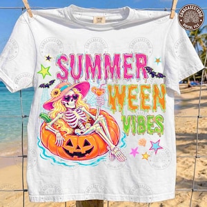 May include: A white t-shirt featuring a graphic of a skeleton wearing a pink sun hat and heart-shaped sunglasses, lounging in a jack-o'-lantern. The text reads "SUMMER WEEN VIBES" in a dripping, colorful font, surrounded by bats and stars.