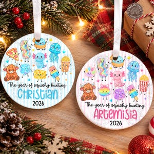 Può includere: Due ornamenti rotondi in ceramica con nastri bianchi. L'ornamento a sinistra ha strisce blu, il testo "The year of squishy hunting CHRISTIAN 2026" e personaggi. Quello a destra ha strisce rosa, il testo "The year of squishy hunting ARTEMISIA 2026" e personaggi.