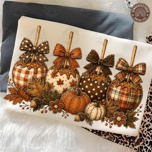 May include: A white t-shirt featuring a graphic print of four caramel apples with decorative bows, each with a unique pattern including gingham, maple leaves, polka dots, and plaid. The design includes two pumpkins, acorns, and autumn leaves.
