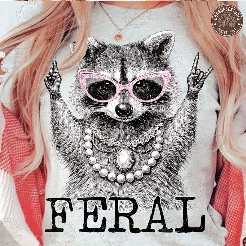 May include: A light gray t-shirt featuring a detailed illustration of a raccoon wearing pink cat-eye glasses and a pearl necklace. The word "FERAL" is printed in bold black letters at the bottom of the design.