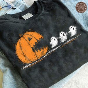 Pode incluir: Camiseta preta com um design de Halloween. Uma abóbora laranja com uma boca irregular está à esquerda, com três fantasmas brancos a segui-la numa linha diagonal. O design tem um estilo vintage desenhado à mão.