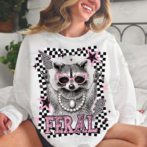 May include: White sweatshirt featuring a raccoon wearing pink sunglasses and a pearl necklace. The raccoon is surrounded by pink stars, lightning bolts, and a black and white checkered border. The word "FERAL" is printed in pink.