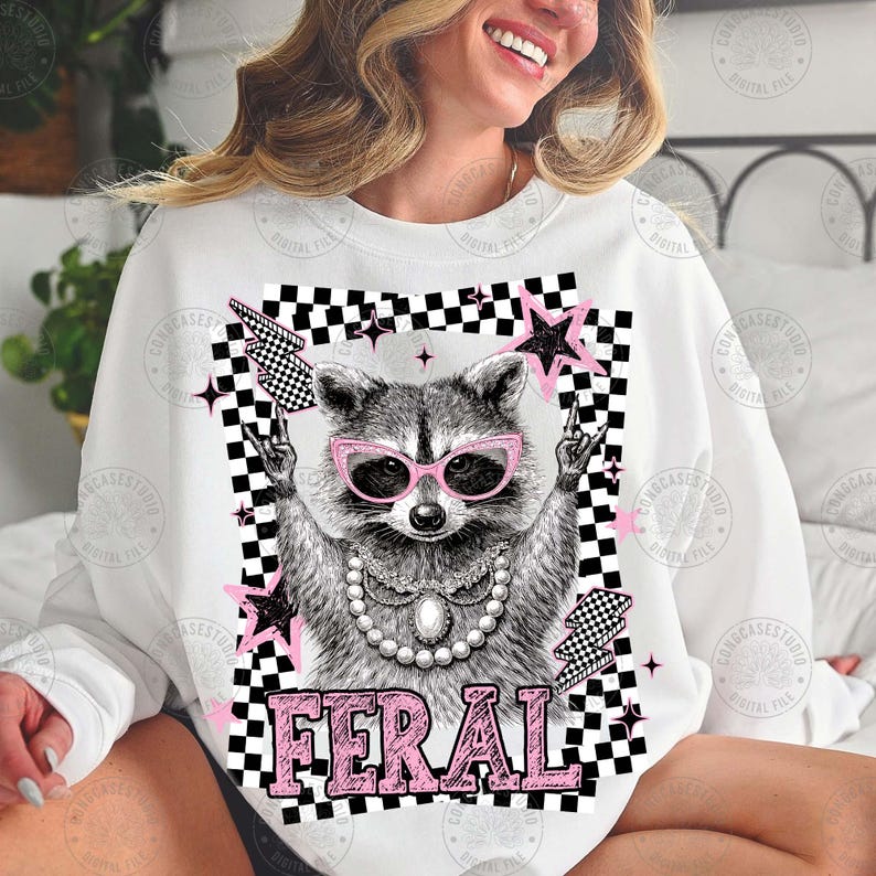 May include: White sweatshirt featuring a raccoon wearing pink sunglasses and a pearl necklace. The raccoon is surrounded by pink stars, lightning bolts, and a black and white checkered border. The word "FERAL" is printed in pink.