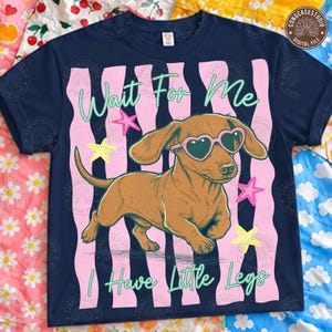 Op de afbeelding: Marineblauw t-shirt met een bruine teckel met een hartvormige zonnebril. Het shirt heeft de tekst "Wait For Me, I Have Little Legs" met roze en gele steraccenten en roze verticale strepen.