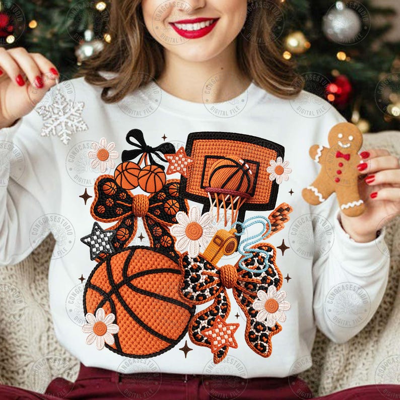 May include: White long-sleeved sweatshirt with a basketball-themed design. The design includes basketballs, a hoop, bows, and flowers. A gingerbread man is held in the hand. The design is in shades of orange, black, and white.