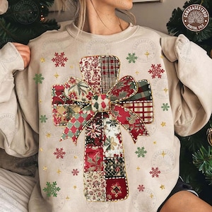 May include: Beige sweatshirt featuring a patchwork cross design with a bow. The cross is made of various patterned fabrics in red, green, and white. Gold snowflakes and stars are scattered around the cross. The sweatshirt has long sleeves.