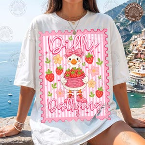 Könnte beinhalten: Weißes T-Shirt mit einer rosa-weißen Grafik. Die Grafik zeigt eine Cartoon-Gans, die einen Korb mit Erdbeeren trägt, umgeben von Blumen und Erdbeeren. Der Text "Dilly Dallying" ist ebenfalls vorhanden.