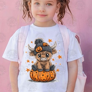 May include: White t-shirt featuring a cartoon Highland cow wearing a black beanie with pumpkin accents, sitting on an orange pumpkin. The design includes orange stars and a speckled background, perfect for Halloween.