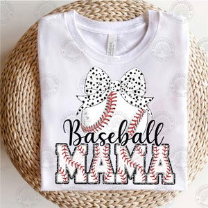 May include: White t-shirt with a black and white polka dot bow on top of a baseball. The text "Baseball Mama" is written in black with red stitching around the letters.