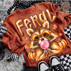 May include: A rust-colored crewneck sweatshirt featuring a graphic of a raccoon wearing orange-rimmed glasses and blowing a pink bubblegum bubble. The text FERAL is printed above the raccoon, surrounded by orange autumn leaves and pumpkins.
