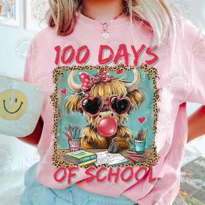May include: A pink t-shirt with the words "100 Days of School". The design includes a cartoon Highland cow wearing heart-shaped sunglasses and blowing a bubble. School supplies surround the cow.