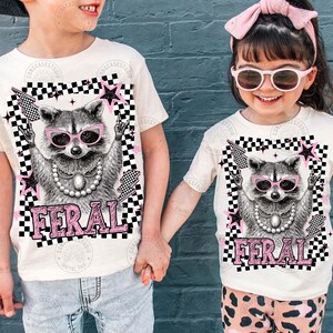 May include: Off-white t-shirts featuring a raccoon wearing pink sunglasses and a pearl necklace. The raccoon graphic is set within a black and white checkered frame with pink stars and lightning bolts. The word "FERAL" is printed in pink.