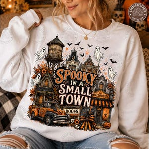 May include: A white long-sleeve sweatshirt featuring a graphic print of a small town with a water tower, houses, and a vintage truck. The text reads "SPOOKY IN A SMALL TOWN" and "90048". The design includes ghosts, bats, and orange autumn foliage.