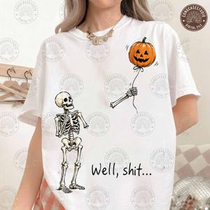 May include: A white t-shirt featuring a graphic of a skeleton standing and looking up at a floating orange jack-o'-lantern pumpkin balloon held by a detached skeletal arm. The text "Well, shit..." is printed in black below the skeleton.