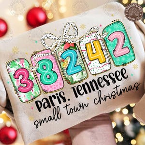 May include: A beige sweatshirt featuring a festive design with the numbers 38242 in colorful tags. The tags are adorned with Christmas-themed patterns and tied with a spotted bow. The text "Paris. Tennessee small town christmas" is printed below.